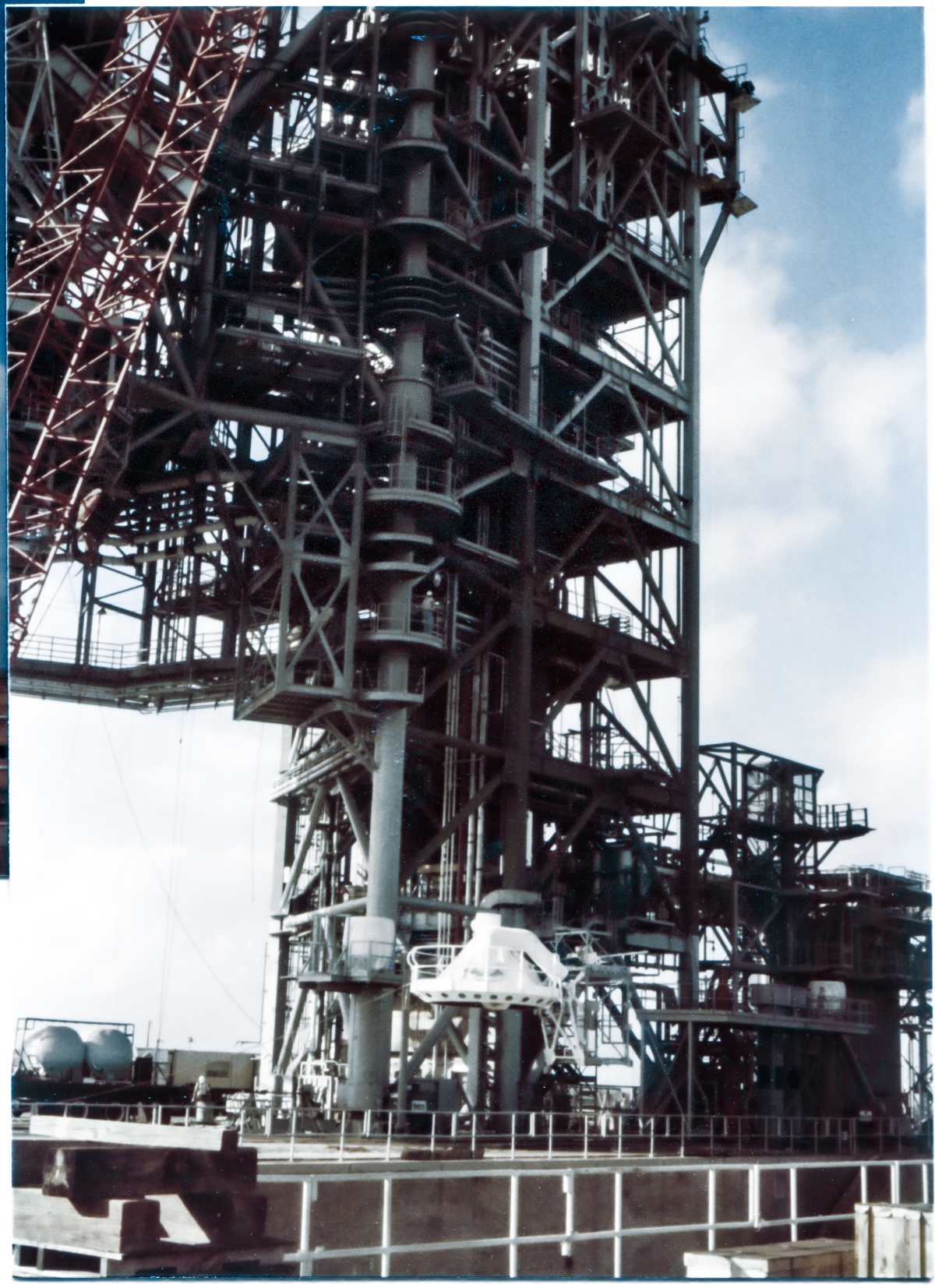 Image 118. With the Fixed Service Structure at Space Shuttle Launch Complex 39-B, Kennedy Space Center, Florida, as backdrop directly behind it, the white flying-saucer-looking ”Beanie Cap” of the External Tank Gaseous Oxygen Vent System Tip Assembly can be seen as it is guided at low elevation over the open (and perforce unoccupied) gap of the Flame Trench by the crane operator to a position where it will then be lifted directly upward to its final elevation nearly 250 feet above the Pad Deck, and then boomed left a little more to where it will be attached by Ivey Steel's Union Ironworkers to the end of the GOX Arm, which is attached to the FSS out of frame above, and not visible in this photograph. Photo by James MacLaren.